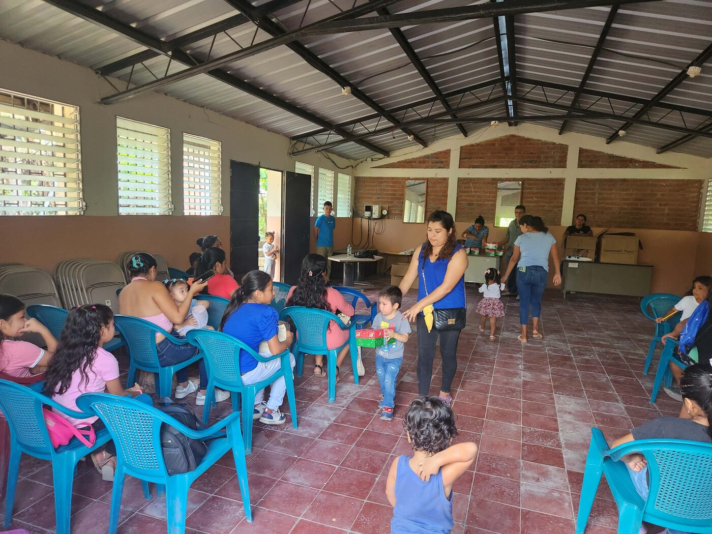 A volunteer distributes shoebox gifts to children while others wait nearby in an open-air pavilion in El Salvador